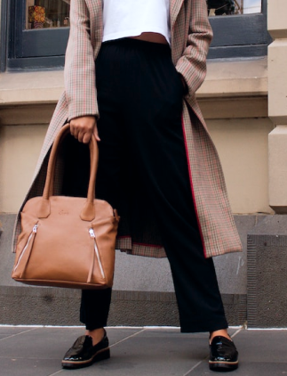 Cintura, pernas e pés de modelo feminina vestindo sobretudo de cor predominante bege com detalhes em tom vermelho e escuro, com calça e par de sapatos na cor preta, segurando bolsa na cor bege, com fundo de prédio.